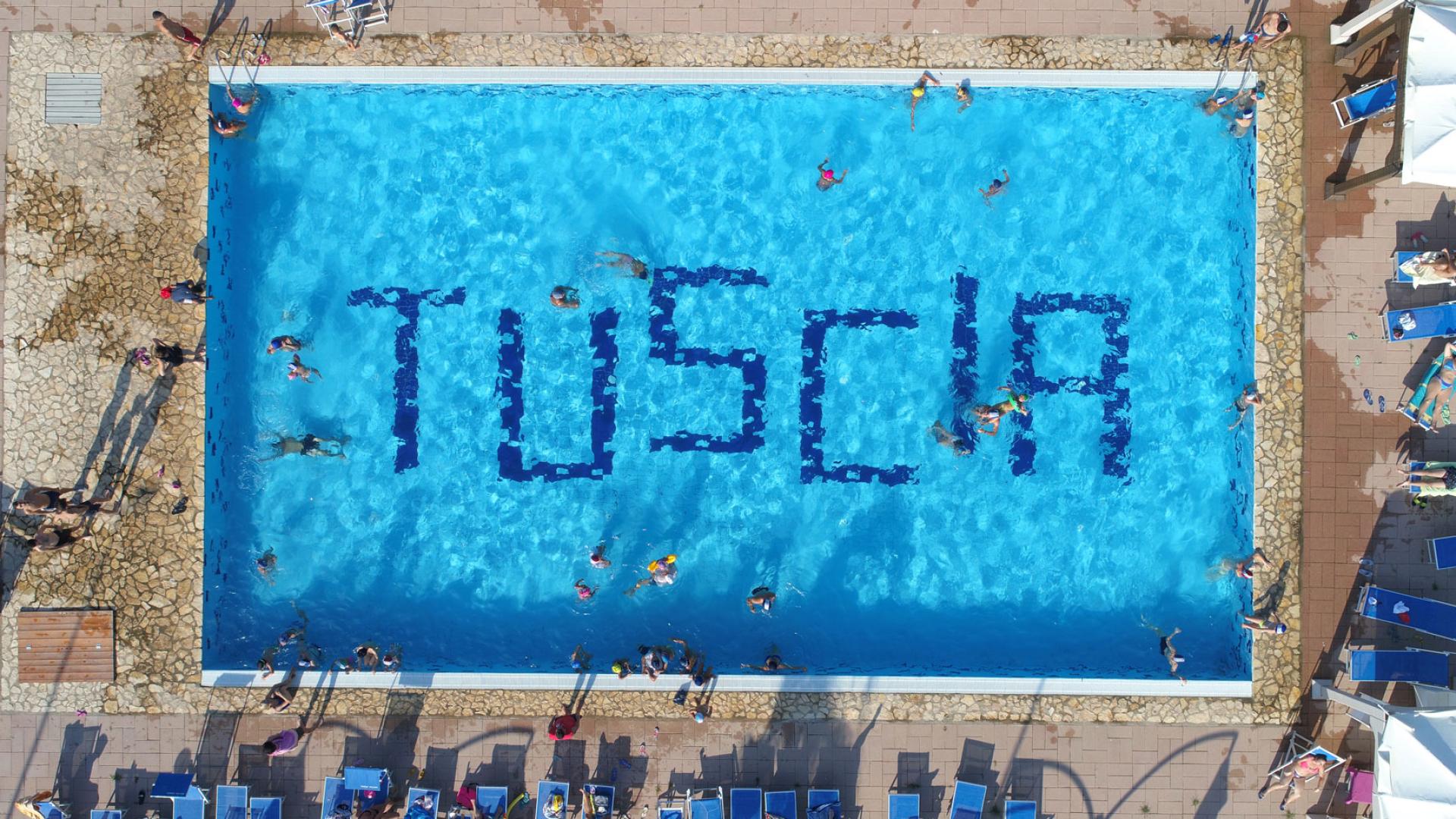 Piscina con scritta 'TUSCIA' vista dall'alto, persone nuotano e si rilassano.