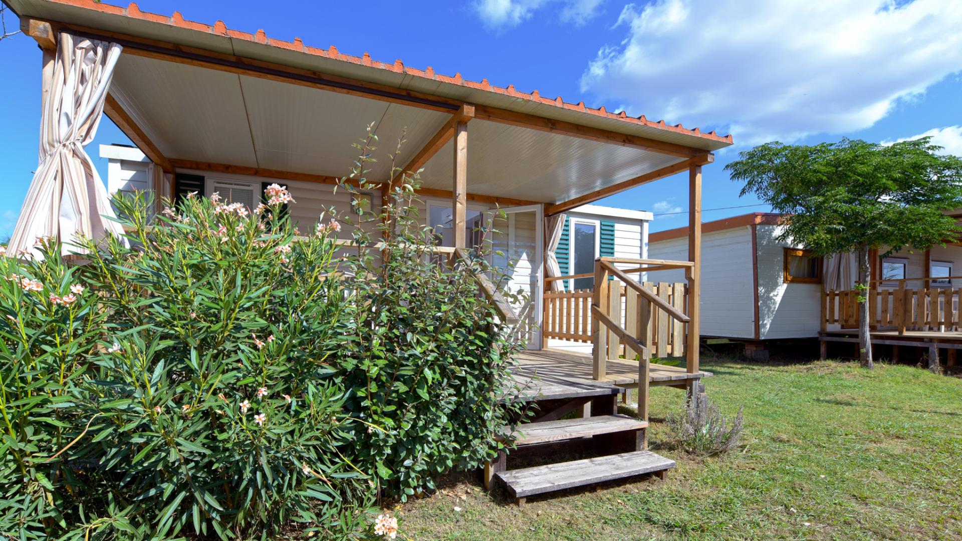 Casetta mobile con veranda in legno, circondata da piante e cielo sereno.