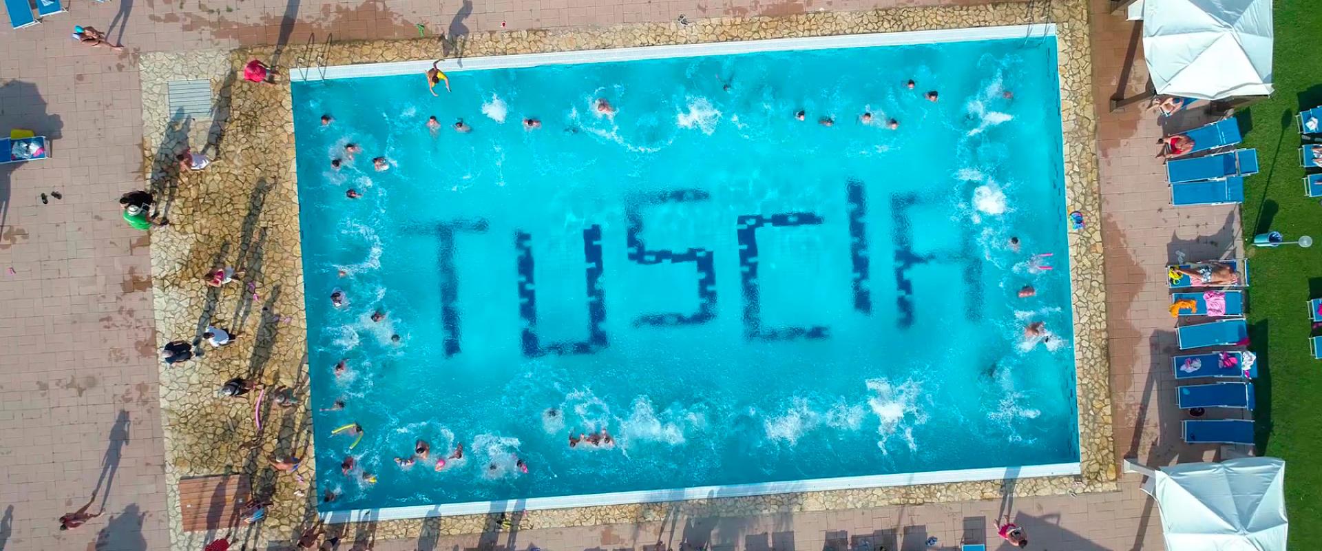 Piscina affollata con lettini e ombrelloni, vista dall'alto, scritta 'TUSCIA' sul fondo.