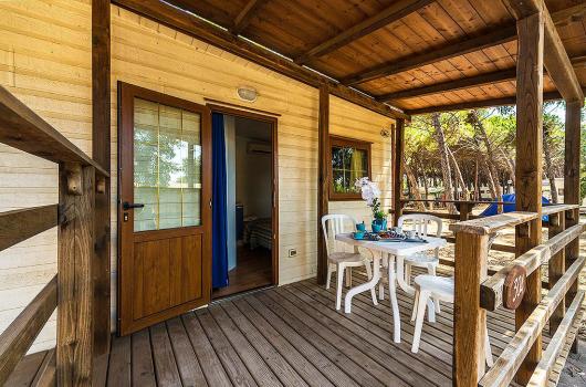 Veranda in legno con tavolo, sedie bianche e vista su un bosco.