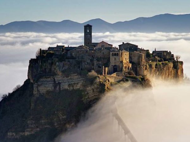 Città medievale su una collina avvolta dalla nebbia, con montagne sullo sfondo.