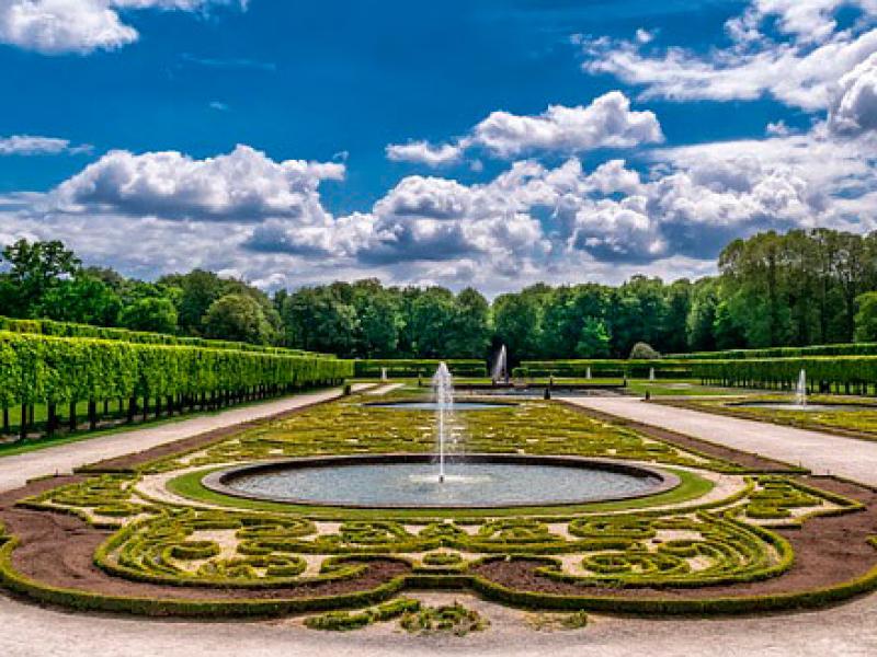 Giardino formale con siepi curate, fontana centrale e cielo nuvoloso.